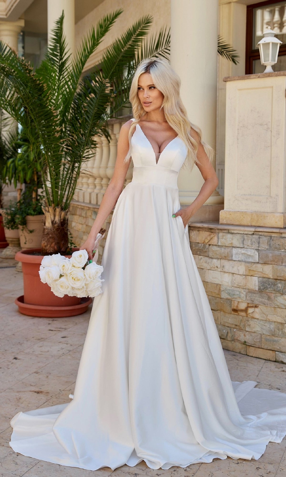 A woman in the Tina Holly BA269W long white satin bridal gown with a deep v-neck holds a bouquet of white roses outdoors near potted plants and stone columns.